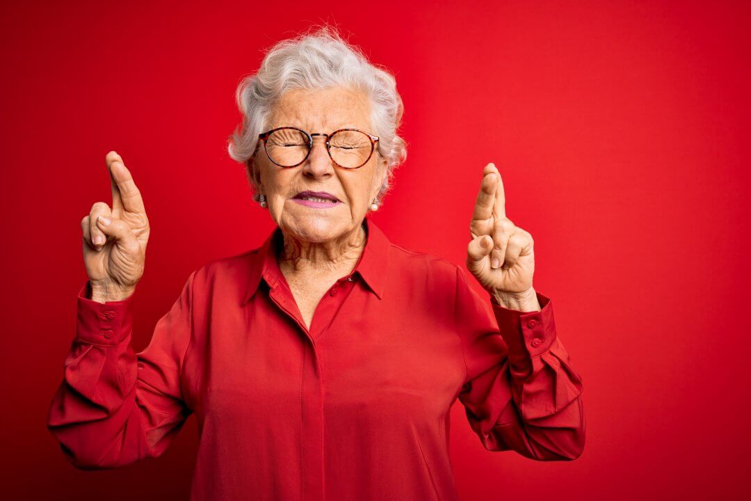 Senior beautiful grey-haired woman wearing casual shirt and glasses over red background gesturing finger crossed smiling with hope and eyes closed. Luck and superstitious concept.
